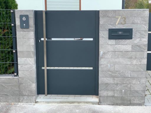 Moderne Eingangstür aus dunklem Metall mit grauem Steinmauerwerk und Hausnummer 73.